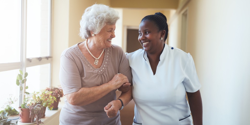 Eine Seniorin und eine Krankenpflegerin gehen einen Flur entlang. Die Pflegerin stuetzt die Seniorin. Beide unterhalten sich und Laecheln.